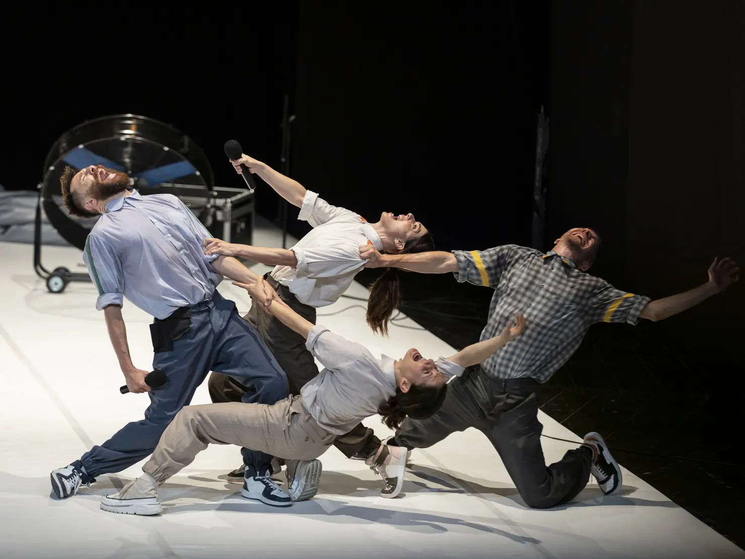 Vier Tänzer*innen in dynamischen Posen auf einer offenen Bühnenfläche. Zwei Tänzer*innen halten Mikrofone, während sie interagieren. Die Atmosphäre ist energiegeladen und performativ.