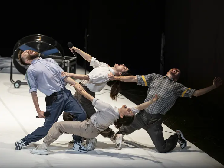 Vier Tänzer*innen in dynamischen Posen auf einer offenen Bühnenfläche. Zwei Tänzer*innen halten Mikrofone, während sie interagieren. Die Atmosphäre ist energiegeladen und performativ.