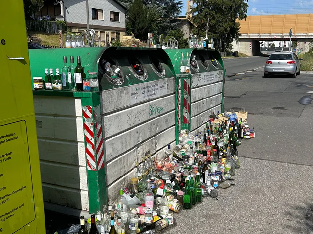 Zwei Altglascontainer an einer Straße mit zahlreichen Glasflaschen und Gläsern, die auf dem Boden davor stehen. Klick öffnet die Meldung.