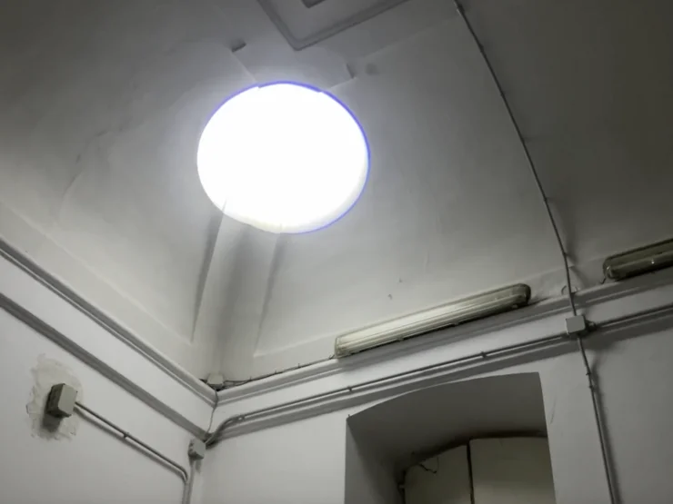 Rundes Oberlicht in einer hohen, weißen Raumdecke mit sichtbaren Kabeln und Leuchtstoffröhre an der Wand