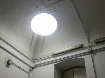Rundes Oberlicht in einer hohen, weißen Raumdecke mit sichtbaren Kabeln und Leuchtstoffröhre an der Wand