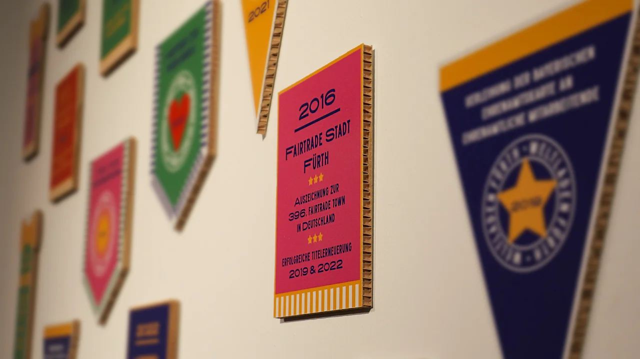 Fairtrade-Auszeichnungen hängen an der Wand, eine davon im Fokus mit dem Text "2016 Fairtrade Stadt Fürth, Auszeichnung zur 396. Fairtrade Town in Deutschland, Erfolgreiche Titelerneuerung 2019 & 2022. Klick öffnet Bild in größerer Ansicht.