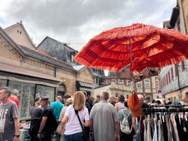 Menschenmenge auf einem Markt mit einem großen roten Sonnenschirm und einem Kleiderständer im Vordergrund. Klick öffnet die Meldung.