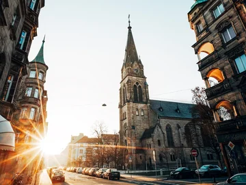 Die Paulskirche in der Südstadt steht an einer Straße, die von Altbauten gesäumt ist, mit tiefstehender Sonne, die Lichtstrahlen über den Gehweg wirft.