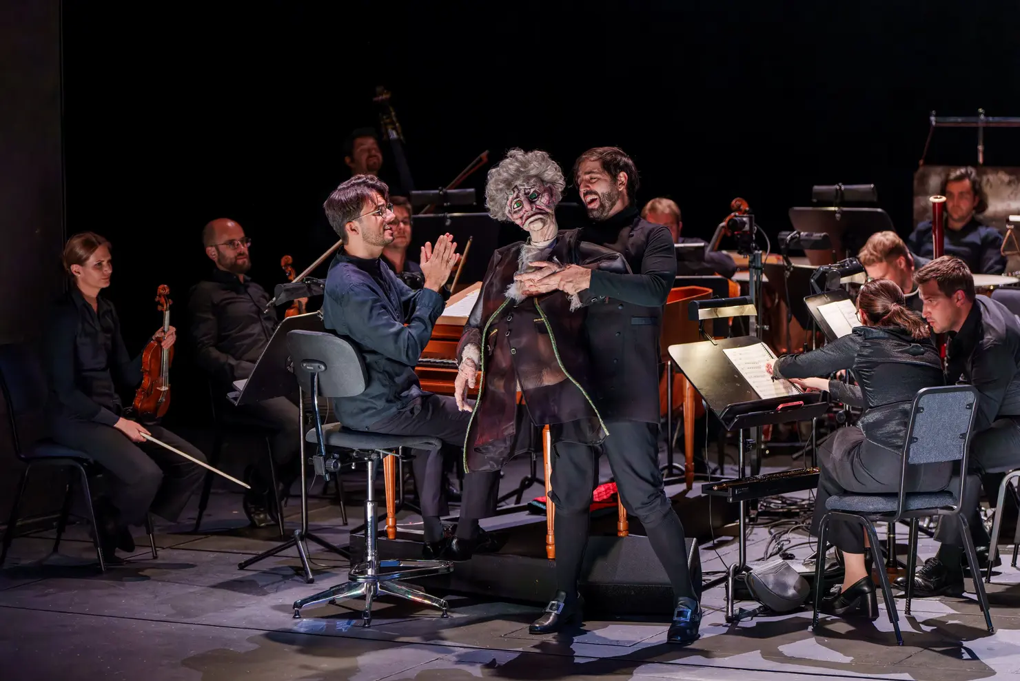 Ein Orchester mit Musiker*innen in schwarzer Kleidung auf der Bühne. Im Vordergrund steht ein Puppenspieler mit einer grotesken Puppe, die eine dramatische Pose einnimmt.