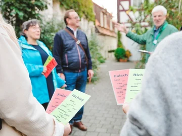 Personen halten grüne und rote Abstimmungskarten mit den Aufschriften 'Richtig' und 'Falsch', im Hintergrund steht ein Stadtführer auf einem gepflasterten Weg.