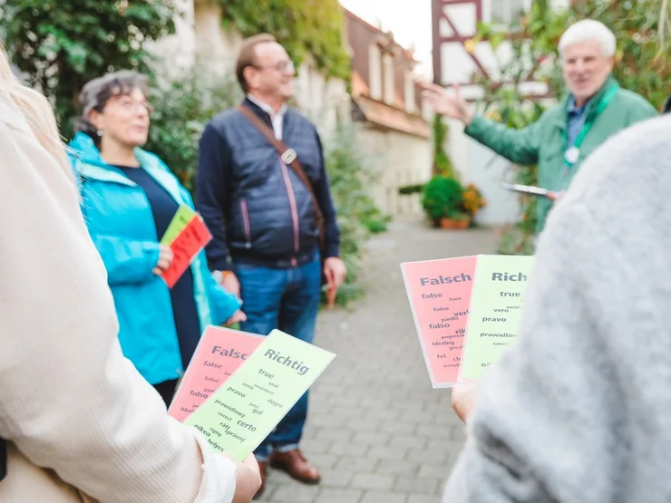 Personen halten grüne und rote Abstimmungskarten mit den Aufschriften 'Richtig' und 'Falsch', im Hintergrund steht ein Stadtführer auf einem gepflasterten Weg.