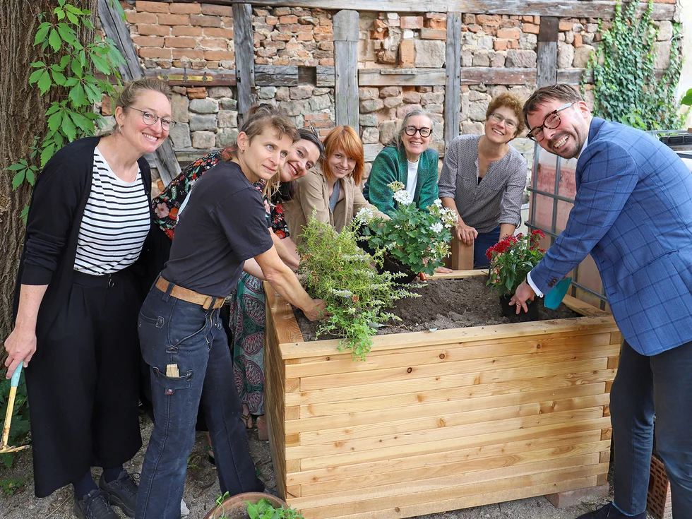 Gruppe von sechs Personen um ein mit Erde und Pflanzen gefülltes Hochbeet aus Holz in einem Innenhof mit Backsteinwand.