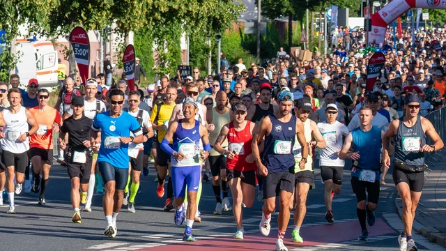 Viele Läuferinnen und Läufer starten bei einem Marathonlauf auf einer von Bäumen gesäumten Straße unter blauem Himmel. Klick öffnet die Meldung.