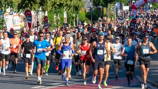 Viele Läuferinnen und Läufer starten bei einem Marathonlauf auf einer von Bäumen gesäumten Straße unter blauem Himmel. Klick öffnet die Meldung.
