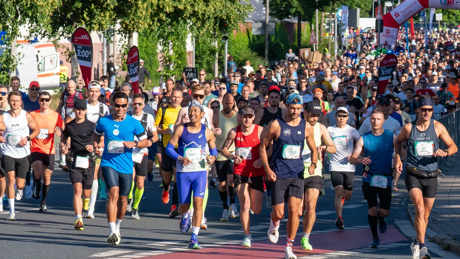 Viele Läuferinnen und Läufer starten bei einem Marathonlauf auf einer von Bäumen gesäumten Straße unter blauem Himmel.