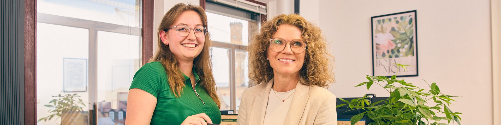 Zwei Frauen stehen in einem Büro vor einem Schreibtisch und betrachten gemeinsam ein Tablet.
