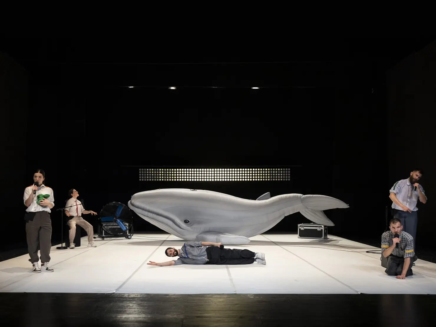 Theateraufführung mit einer großen Walskulptur und mehreren Schauspieler*innen in unterschiedlichen Posen, darunter ein liegender Schauspieler und halten oder sitzen an einem Mikrofon.