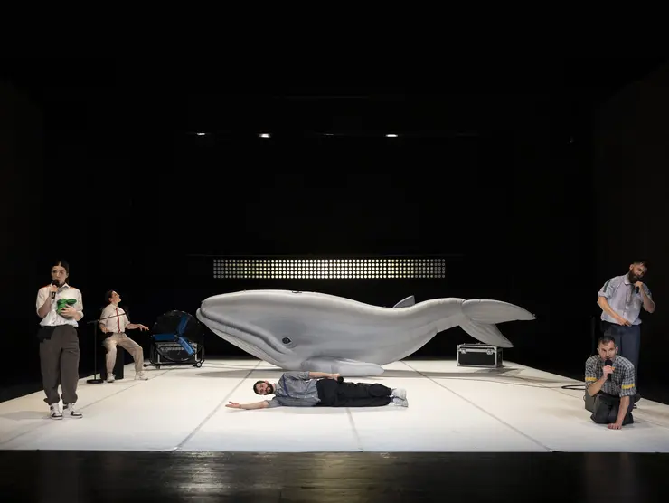 Theateraufführung mit einer großen Walskulptur und mehreren Schauspieler*innen in unterschiedlichen Posen, darunter ein liegender Schauspieler und halten oder sitzen an einem Mikrofon.