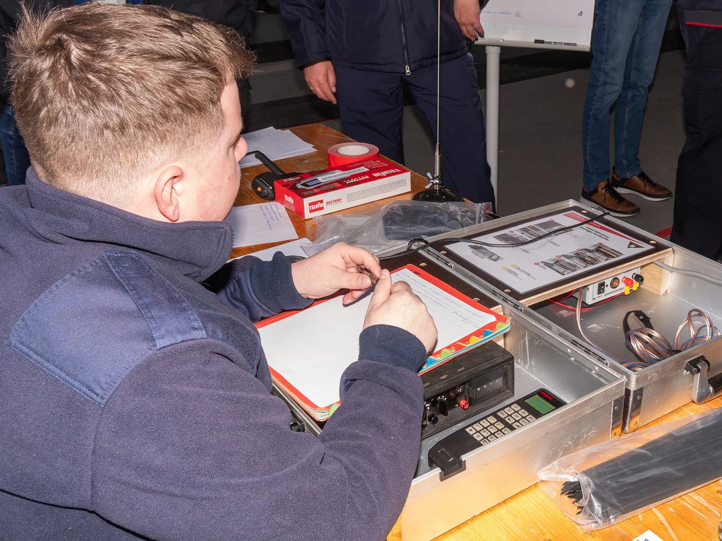 Person sitzt an einem Tisch mit geöffnetem Metallkoffer, in dem sich ein Funkgerät befindet.
