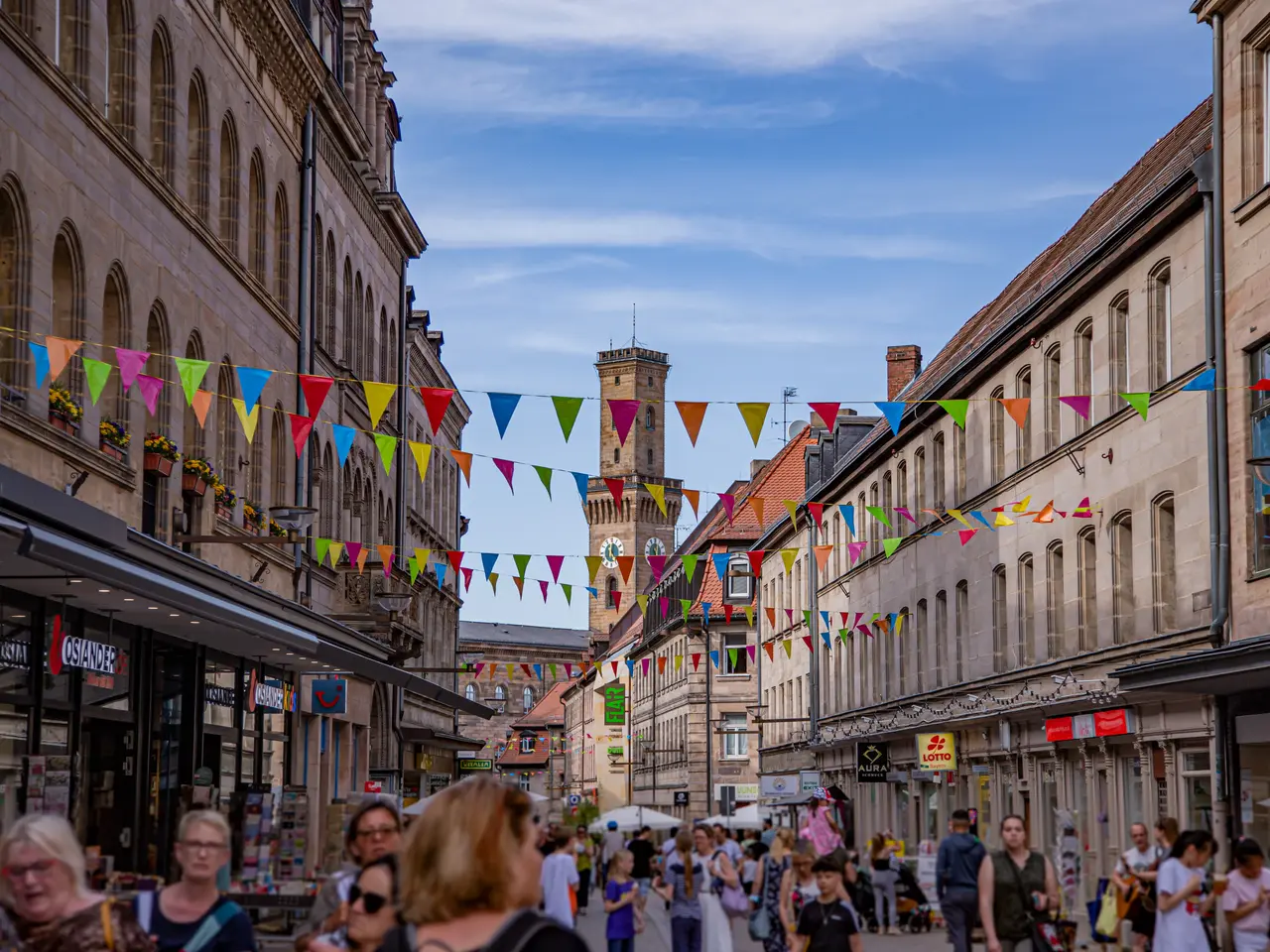 Die belebte Fußgängerzone mit dem Rathausturm im Hintergrund. Bunte Wimpel sind über die Straße gepannt.