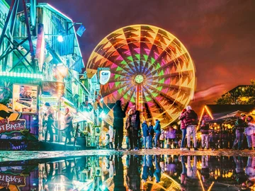 Bunt beleuchtetes Riesenrad auf der Michaelis-Kirchweih mit Spiegelung im Wasser und Menschen davor bei Nacht.
