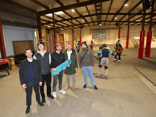 Gruppenfoto der beteiligten Personen in der Halle und skatende Jugendliche im Hintergrund. Klick öffnet die Meldung.