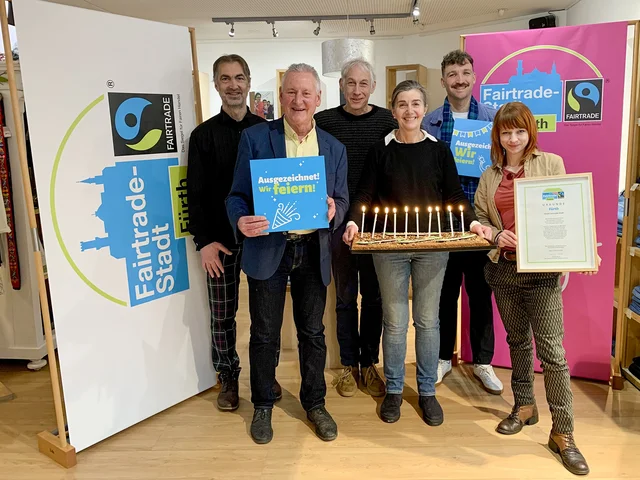 Die genannten Personen stehen im Weltladen vor zwei Fairtrade Town Schildern und halten einen Geburtstagskuchen mit Kerzen in Händen. Klick öffnet die Meldung.