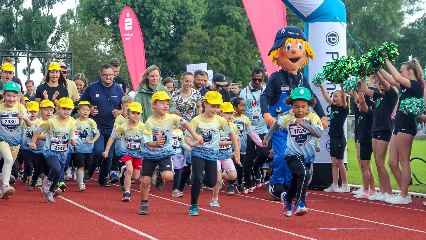Kinder mit gelben Mützen und Laufnummern starten auf einer Laufbahn vor Zuschauern und einer Person in einem bunten Maskottchenkostüm.