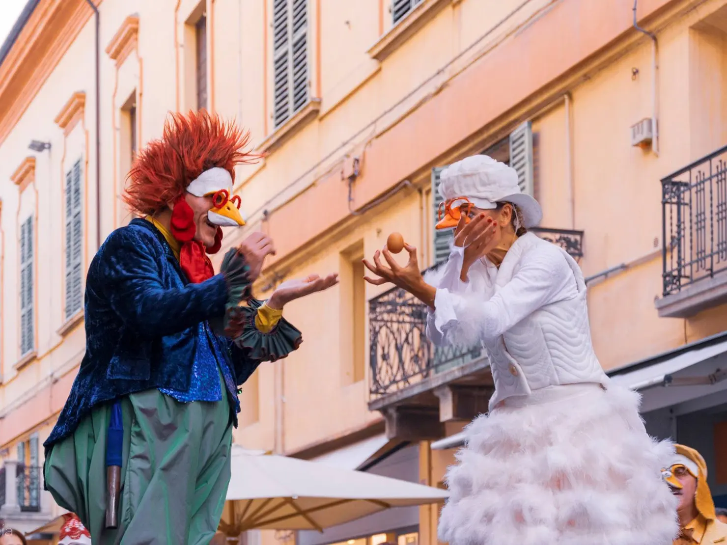 Zwei Personen in bunten Kostümen, einer als Huhn und der andere als Hahn, führen eine humorvolle Darbietung auf einer Straße vor historischen Gebäuden auf.
