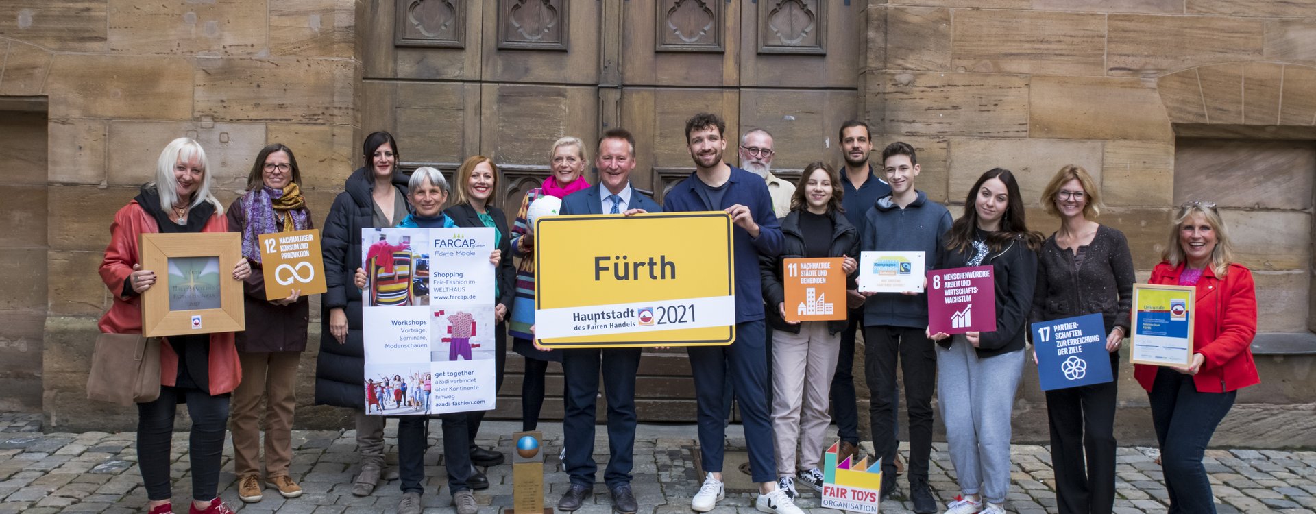 Vertreterinnen und Vertreter der fairen Einrichtungen halten ein gelbes Ortsschild mit der Aufschrift "Fürth. Hauptstadt des Fairen Handels 2021" sowie kleinere Schilder mit den Namen der fairen Fürther Organisationen hoch.