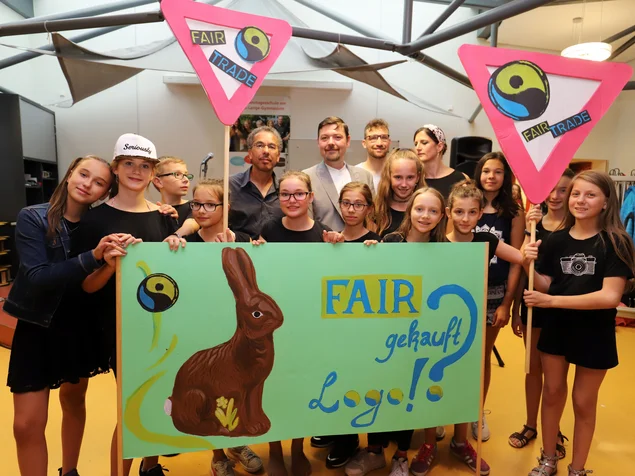 Schülerinnen und Schüler halten ein Schild mit der Aufschrift "Fair gekauft? Logo!", einem Hasenmotiv und Fairtrade-Logo sowie zwei dreieckige Fairtrade-Schilder in die Luft. Klick öffnet Bild in größerer Ansicht.