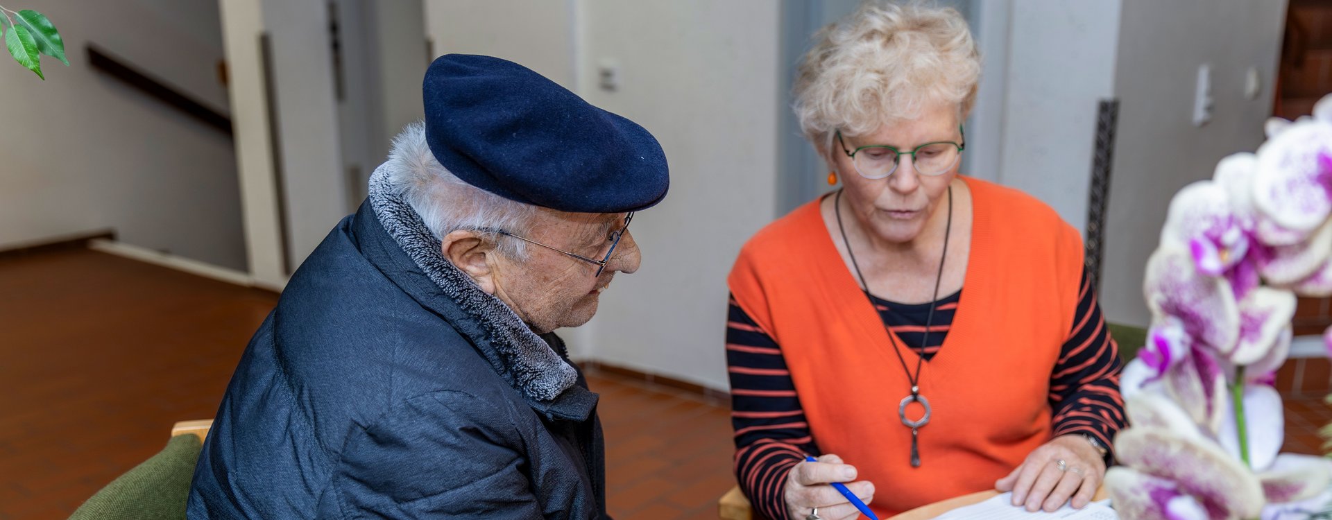 Die Seniorenbeauftragte sitzt mit einem Senioren und einer Frau, die man nur von hinten sitzt an einem Tisch und zeigt ihnen Dokumente.