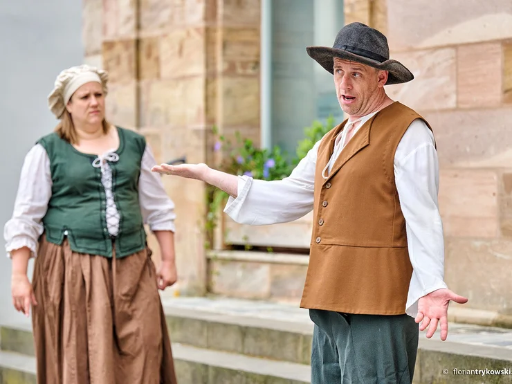 Zwei Schauspieler in historischer Kleidung stehen vor einem Gebäude, einer gestikuliert mit ausgestrecktem Arm, die andere steht im Hintergrund