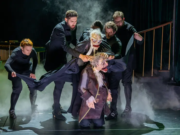 Szenenbild aus einem Theaterstück mit fünf Schauspieler*innen in schwarzer Kleidung, die zwei Puppen mit groteskem Gesicht und grauen Haaren auf der Bühne spielen. Nebel und dunkler Hintergrund schaffen eine dramatische Atmosphäre.