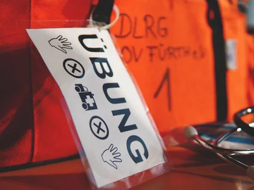 Schild mit der Aufschrift 'ÜBUNG' und Symbolen für Hände, ein Fahrzeug mit Kreuz und weitere Hände vor einem orangefarbenen Hintergrund mit unscharfem Text.