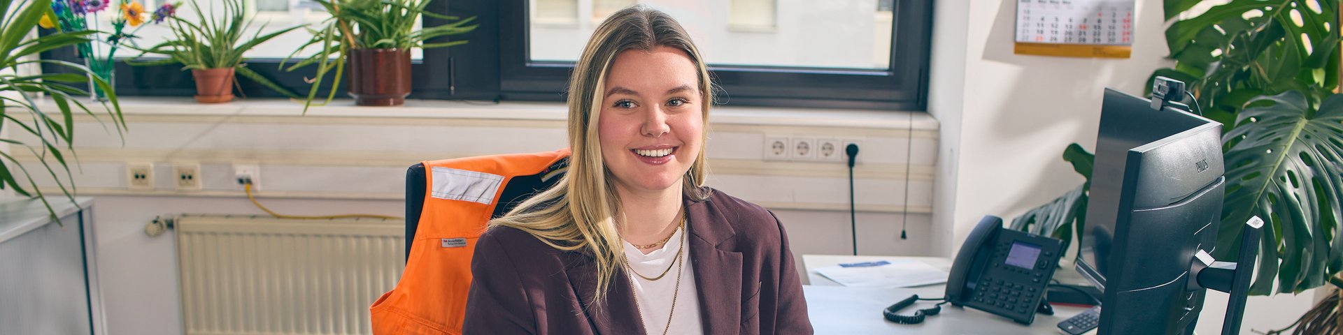 Junge Frau sitzt an einem Schreibtisch mit Warnweste auf dem Bürostuhl. 