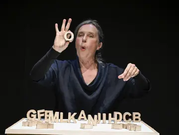 Frau mit grauen Haaren in dunkler Kleidung führt eine Handbewegung aus und hält einen runden Gegenstand. Vor ihr stehen Holzbuchstaben auf einem Tisch.