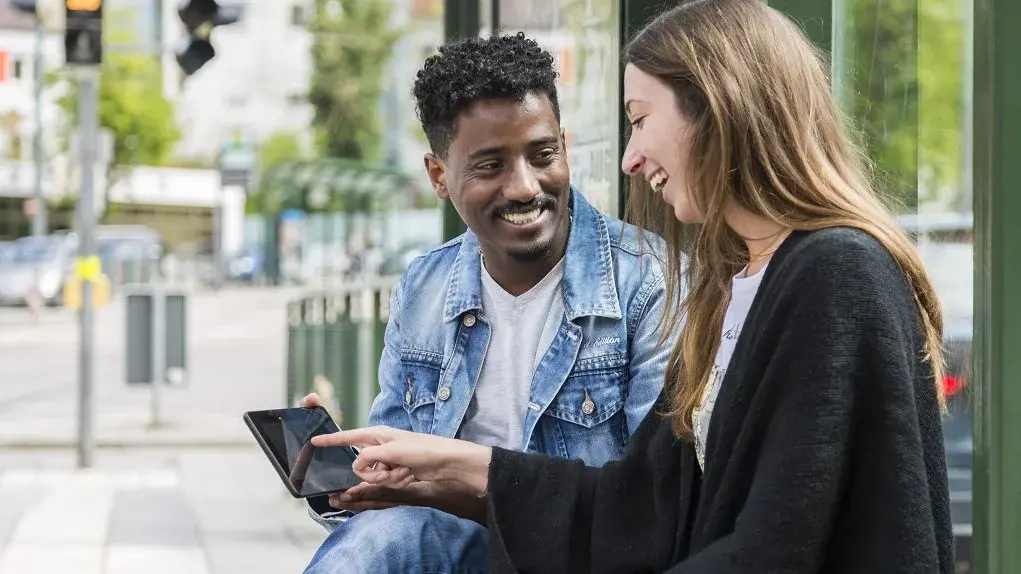 Integreat-App Ein Mann und eine Frau sitzen an einer Haltestelle, sie zeigt auf sein Tablet