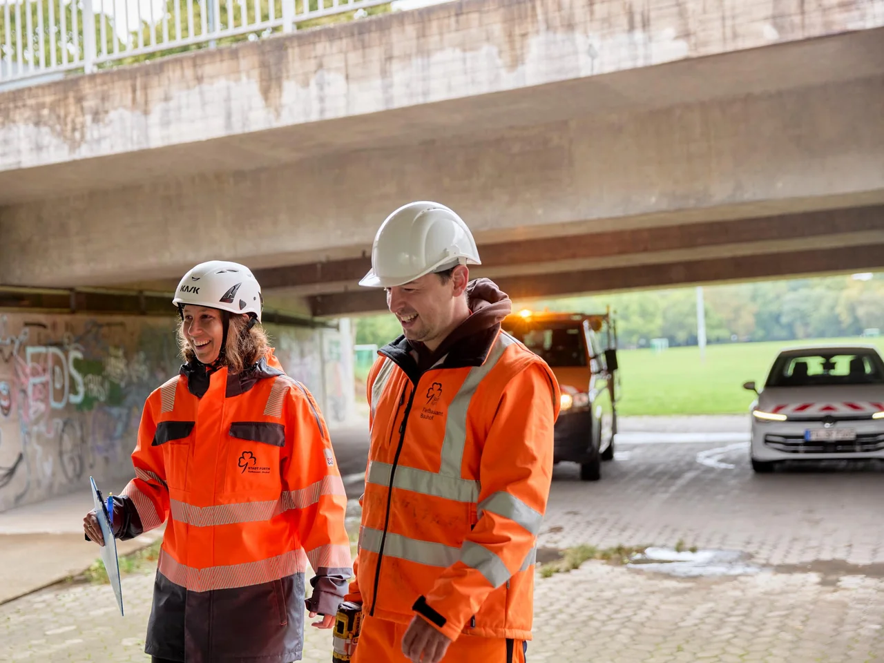 Zwei Personen in orangefarbenen Warnwesten und Schutzhelmen stehen unter einer Brücke, eine hält ein Klemmbrett.
