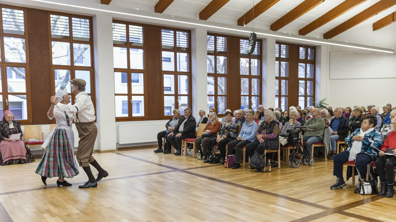 Seniorinnen und Senioren sitzen in einem Saal und schauen einem Paar beim Tanzen zu. Klick öffnet Bild in größerer Ansicht.