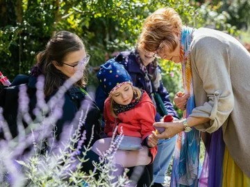 Märchenerzählerin Zorica Otto zeigt einem Kind im Stadtpark etwas in der Hand, umgeben von weiteren Personen und Pflanzen.