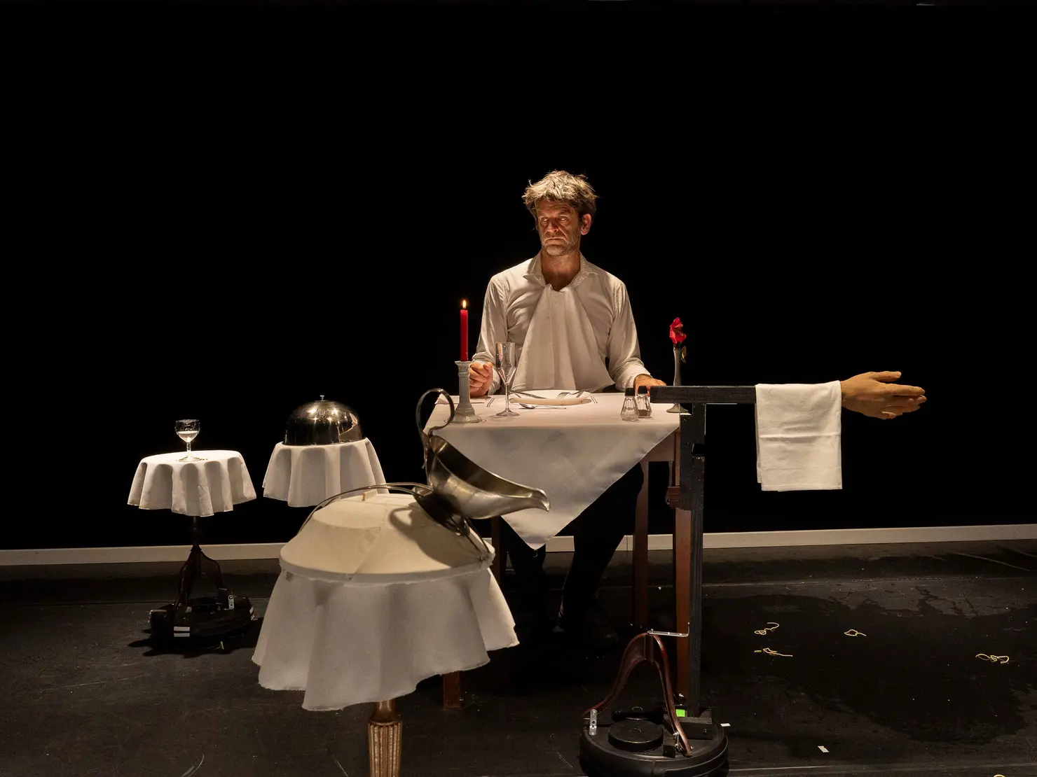 Eine bizarre Inszenierung mit einem Tisch, an dem ein Mensch sitzt. Eine Tischdecke bedeckt den Tisch und eine große Hand ragt unter einem Serviettenring hervor. Der Ausdruck der Person ist neutral. Im Hintergrund stehen zwei kleine Tische mit einem Glas und einer Speiseglocke. Im Vordergrund ein Tisch mit einer Sauciere.