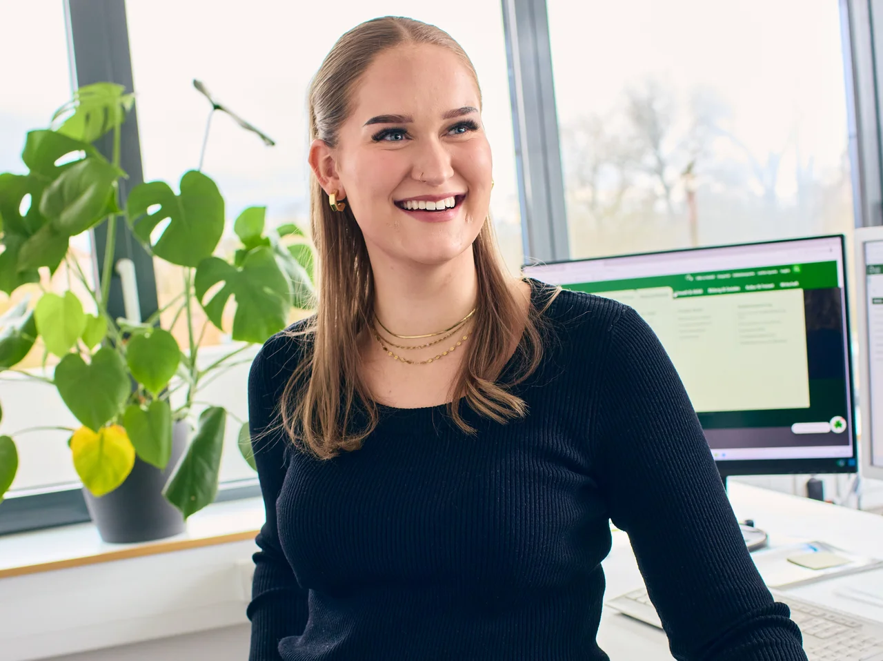 Junge Frau lehnt an einem Schreibtisch in einem Büro und blickt lächelnd. 