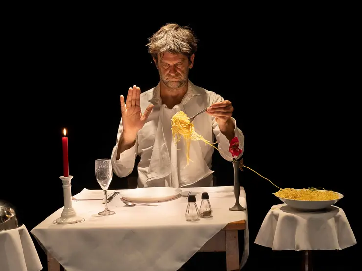 Mann mit grauen Haaren in weißem Hemd an eleganter Tafel. Eine Gabel voller Spaghetti. Die Gabel wird von dem Mann gehalten. Rote Rose und Kerze vorhanden, romantische Atmosphäre.