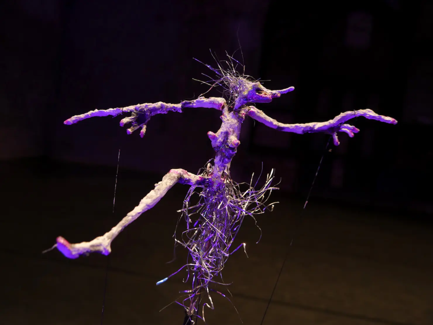 Skulptur eines stilisierten, humanoiden Geschöpfs mit langen Armen und spitzem Gesicht, aus organischen Materialien und Draht, im düsteren Hintergrund beleuchtet.