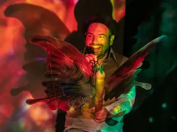 Mann mit großem bunten Schmetterlingskostüm und Mikrofon, vor einer bunten Lichtprojektion stehend und lächelnd.