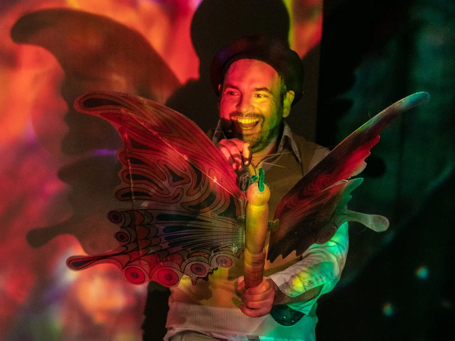 Mann mit großem bunten Schmetterlingskostüm und Mikrofon, vor einer bunten Lichtprojektion stehend und lächelnd.