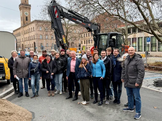 Gruppenbild der Genannten auf dem Königsplatz mit Bagger und Rathaus im Hintergrund. Klick öffnet die Meldung.