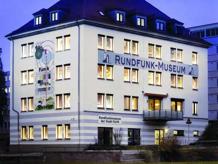 Das dreistöckige, weiße Gebäude des Rundfunkmuseums in der Abenddämmerung mit beleuteten Fenstern. 